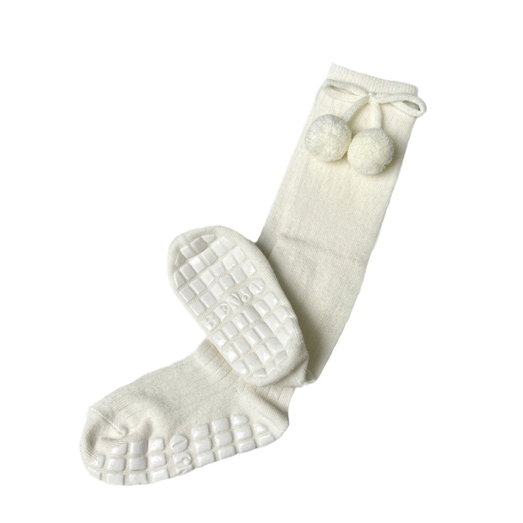 Pair of white knee-high socks with textured rubber grip soles and decorative pom-poms near the top, laid flat against a plain background.